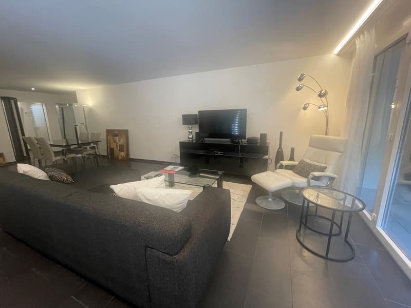 Spacious living room with large windows, modern furniture including a sofa, armchair, coffee table, and TV stand. The room has a minimalist and contemporary design with neutral colors and accents.