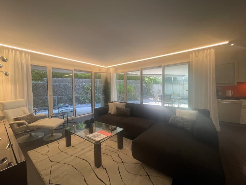 Spacious living room with large windows overlooking a balcony or terrace, comfortable sofa, glass coffee table, and modern lighting fixtures.