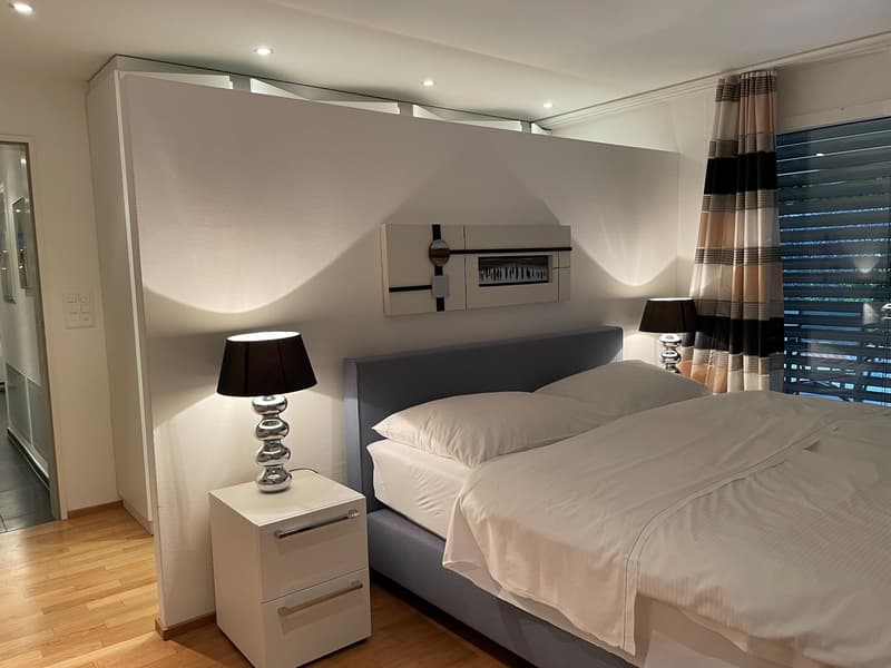 Spacious bedroom with hardwood flooring, gray and white color scheme, large windows, gray upholstered bed frame, nightstands, and decorative lamps.