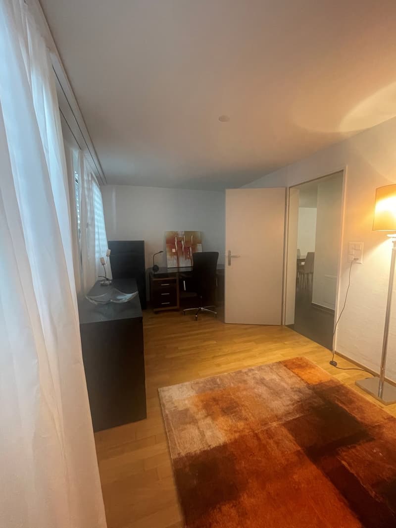 The image shows a bedroom with a hardwood floor, white walls, and large windows. There is a desk with a computer setup, a cabinet, and a rug on the floor. The room appears to be well-lit and cozy.