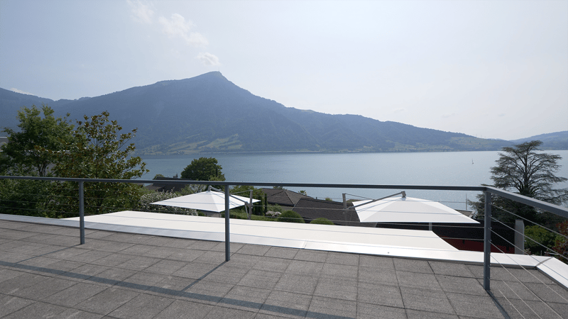 Outdoor terrace with lake view, steel railing, two white umbrellas, tiled floor, trees, mountains in background, calm water