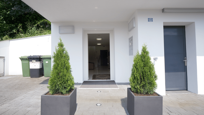 White building, gray door, green trash bins, two potted evergreens, entryway with carpet, number 10 above the door, trash bin area beside the entrance