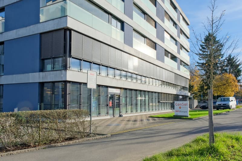 Modern office building, multiple windows, blue exterior walls, large glass doors, shrubs in front