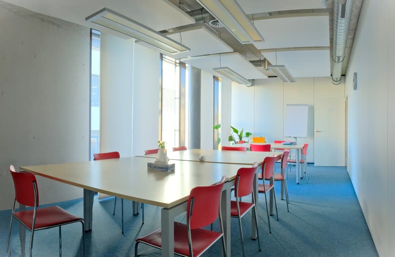 a room with blue carpet, wooden tables, and red chairs, long white walls, white ceiling with lights and pipes, whiteboard, glass windows, white door