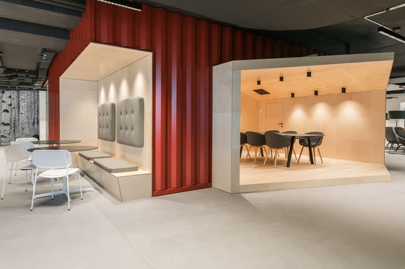 The image shows a modern and stylish office space with a mix of seating areas and meeting rooms. The space features red curtains, white walls, and a combination of light and dark furniture. There is a seating area with tufted gray chairs and a table, as w