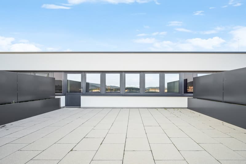 A modern, minimalist building with large windows offering a scenic view of the surrounding landscape. The building has a clean, white exterior with a flat roof and a spacious outdoor terrace or balcony area with black planters or seating elements.