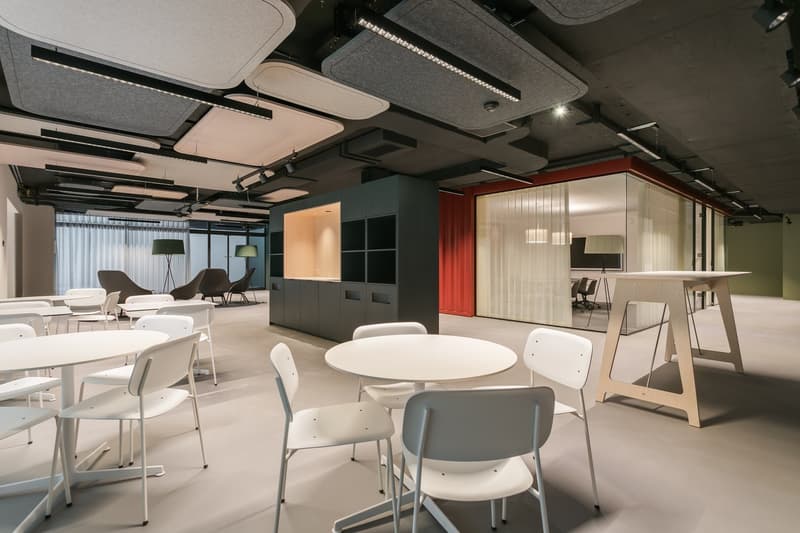 Modern and open office space with round tables, white chairs, and various seating areas. The ceiling has a unique design with lighting fixtures. There are also storage cabinets and a glass partition separating different sections of the space.