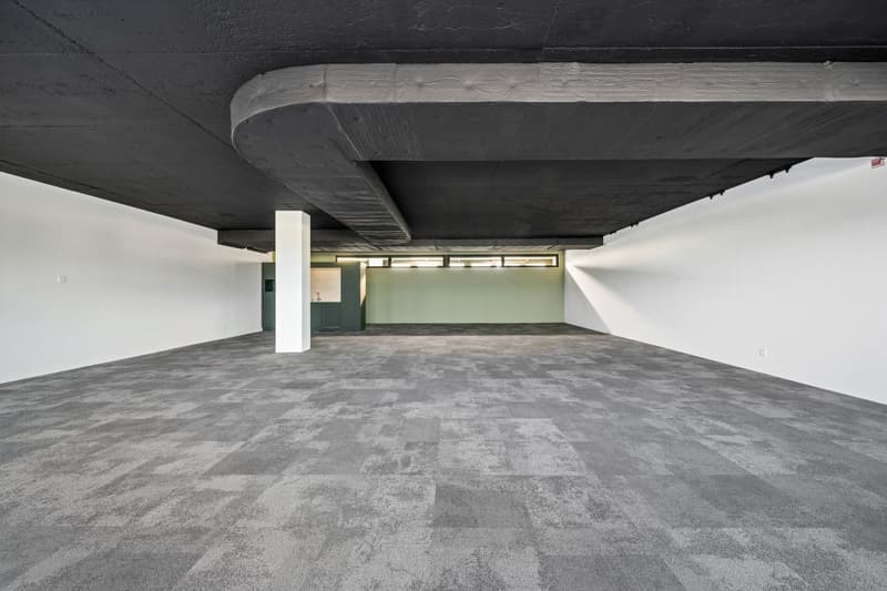 Large open space with concrete floor, white walls, and exposed concrete ceiling with lighting fixtures. The space appears to be a commercial or industrial area, potentially a warehouse or office space.