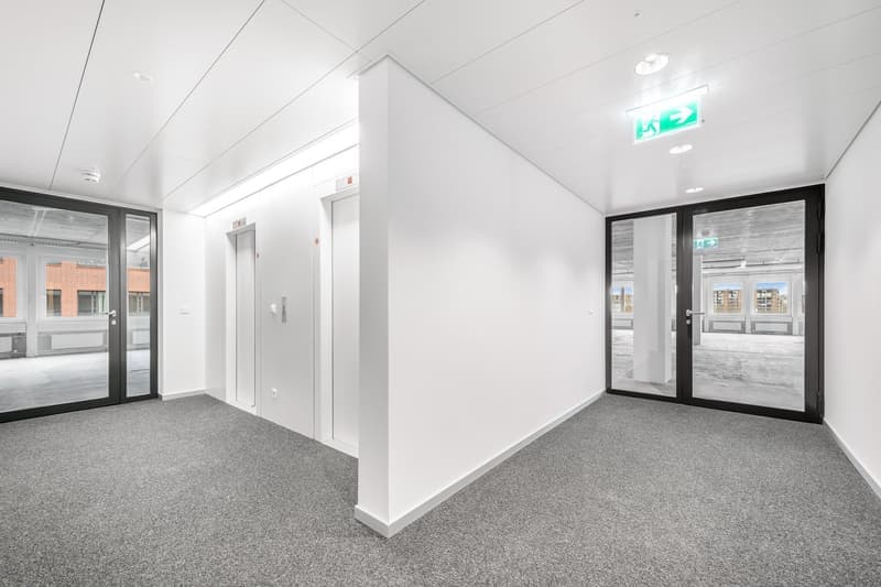 The image shows a spacious, modern office space with white walls, gray carpeted floors, and large glass windows. The room has a high ceiling and appears to be a commercial or business space.