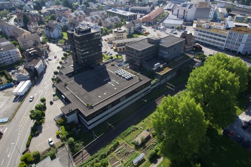 Large building with flat roof, situated on a corner, surrounded by roads, a parking lot, and greenery