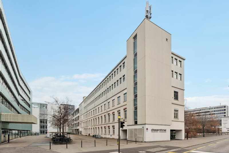 White exterior, multiple floors, rectangular windows, two entrances with text 'dreamtime', antennas on the roof, trees and bicycle stands in front, buildings behind the main building