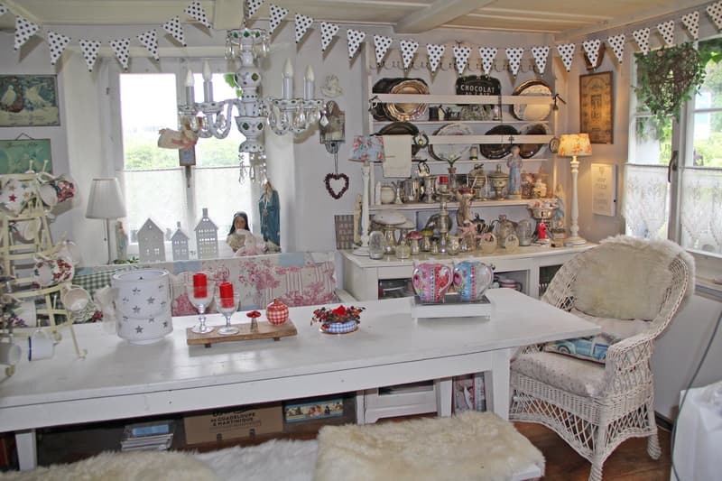 Cozy and eclectic living room with polka dot bunting, vintage decor items, shelves displaying various decorative pieces, a wicker chair, and a table with a dishwasher visible.