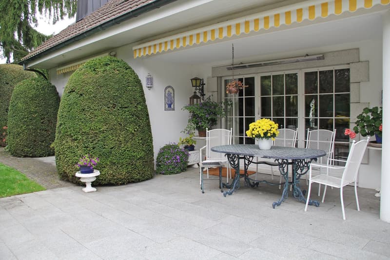 Outdoor dining area with table and chairs, yellow flowers, green hedges, white house, awning
