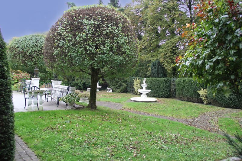A garden with a fountain, trees, shrubs, and a paved walkway. There are also tables and chairs set up for outdoor seating.