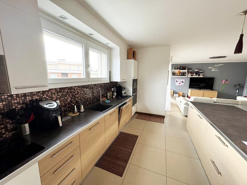 Spacious kitchen with wooden cabinets, black countertop, mosaic tile backsplash, and a dishwasher. The kitchen opens up to a living room area with a TV and other furnishings.