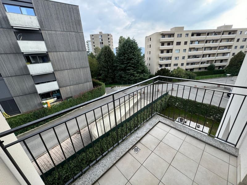 The image shows a balcony or terrace of an apartment building. The balcony has a metal railing and overlooks a landscaped area with trees and other apartment buildings in the background. The building appears to be made of concrete or a similar material wi
