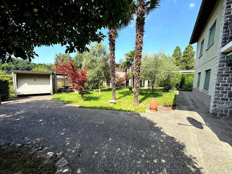 Two-story house with brick exterior, surrounded by lush greenery including palm trees, shrubs, and a grassy lawn. There is a paved driveway or parking area in the foreground, and a small outbuilding or shed visible on the property.