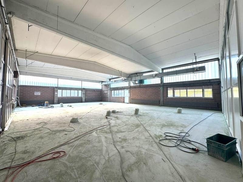 An empty room with high ceiling, metal structures, white walls, and several windows. Wires are lying on the floor and there are barrels and some construction materials.