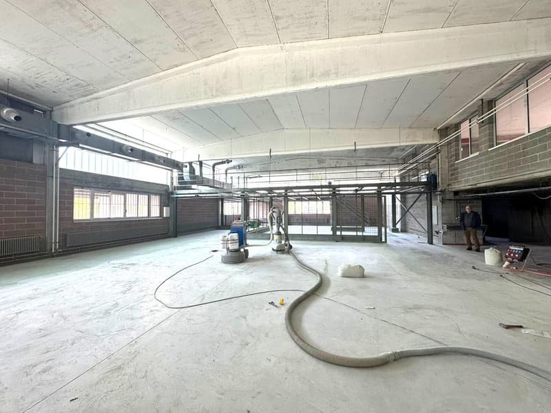 An empty room, concrete floor, large windows, ceiling with multiple metal structures, ventilation systems, a man is standing on the right side of the room