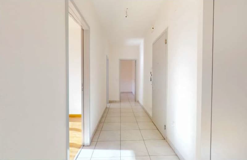 Empty hallway with white tiles and white walls, two doors on either side, one leading to a room with wooden floor.