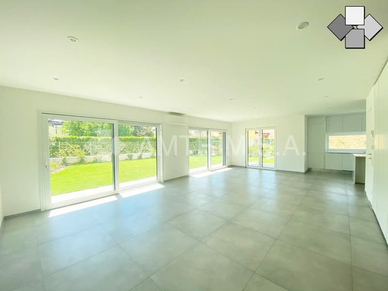 Spacious open-plan living area with large windows overlooking a well-maintained garden, tiled flooring, and a modern kitchen visible in the background.