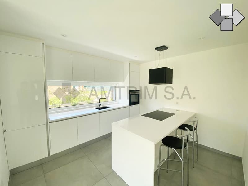 Modern, minimalist kitchen with white cabinets, white countertop, built-in appliances including a dishwasher, and large window providing natural light.
