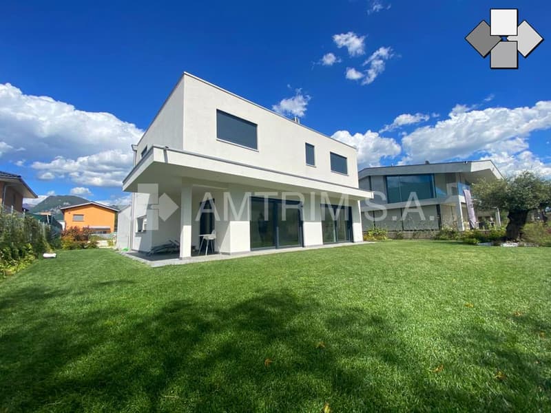 Two-story modern house with a flat roof, large windows, and a garage. The house is surrounded by a well-maintained lawn and garden.