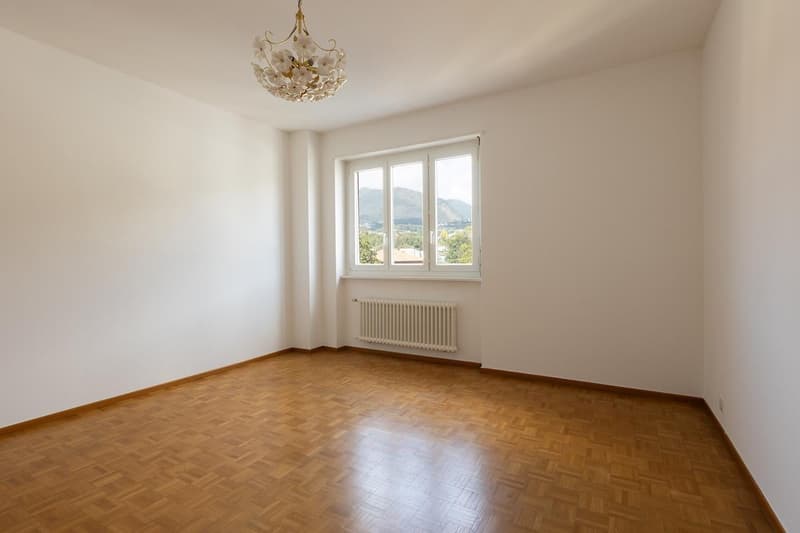 Empty room with wooden floors, white walls, window, and radiator. Chandelier above.