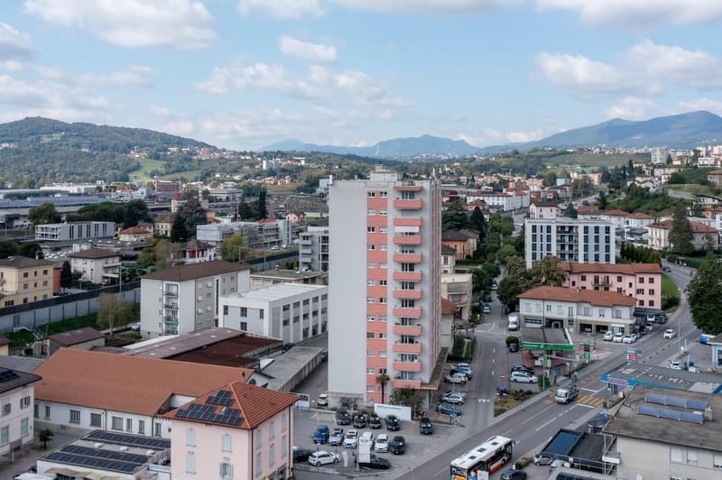 Aerial view of a city with various buildings, mountains in the background, roads, cars parked, solar panels on roofs, streetlights, and a bus.
