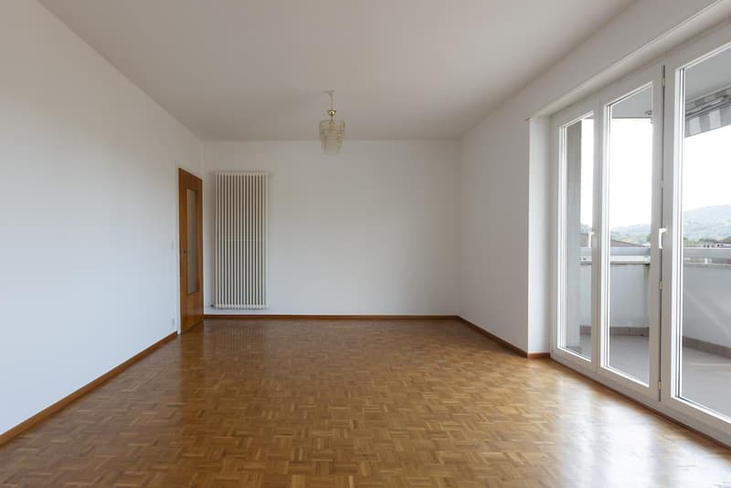 Empty room with wooden parquet flooring, white walls, a door leading to another room, a radiator, large glass doors opening to a balcony with a railing, and a chandelier hanging from the ceiling.