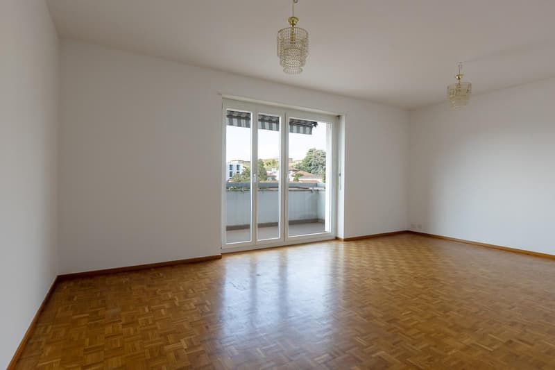 empty room, white walls, wooden floor, sliding glass doors leading to a balcony with a railing, two chandeliers on the ceiling, a view of the outdoors