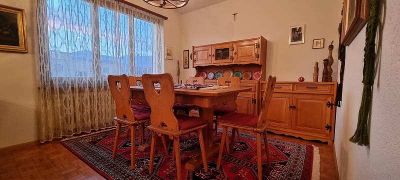 Wooden dining table, chairs, wooden cabinet with plates, curtains, pictures on wall, carpet on the floor