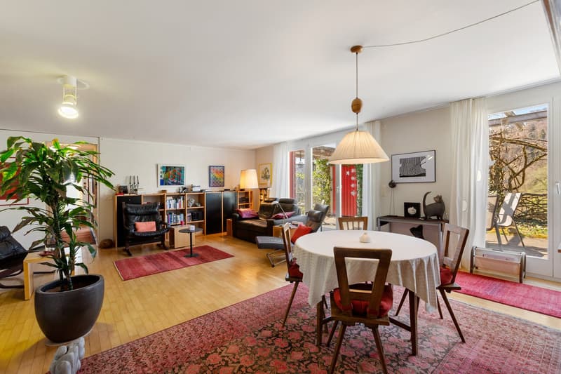 dining area with table and chairs, living area with couch and armchairs, wooden floor, large windows, balcony, potted plant, rug