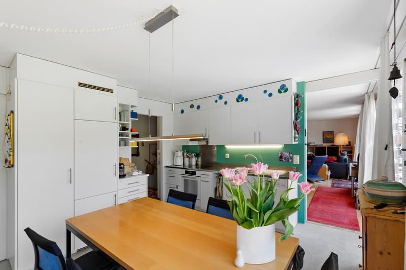 modern kitchen with dining area, white cabinets, wooden table, pink flowers in vase, modern lighting, green wall with blue and green circles
