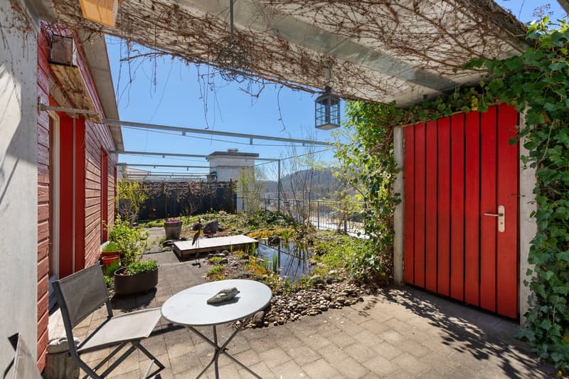 Outdoor area with a table, chair, and a small pond, surrounded by plants and a red gate