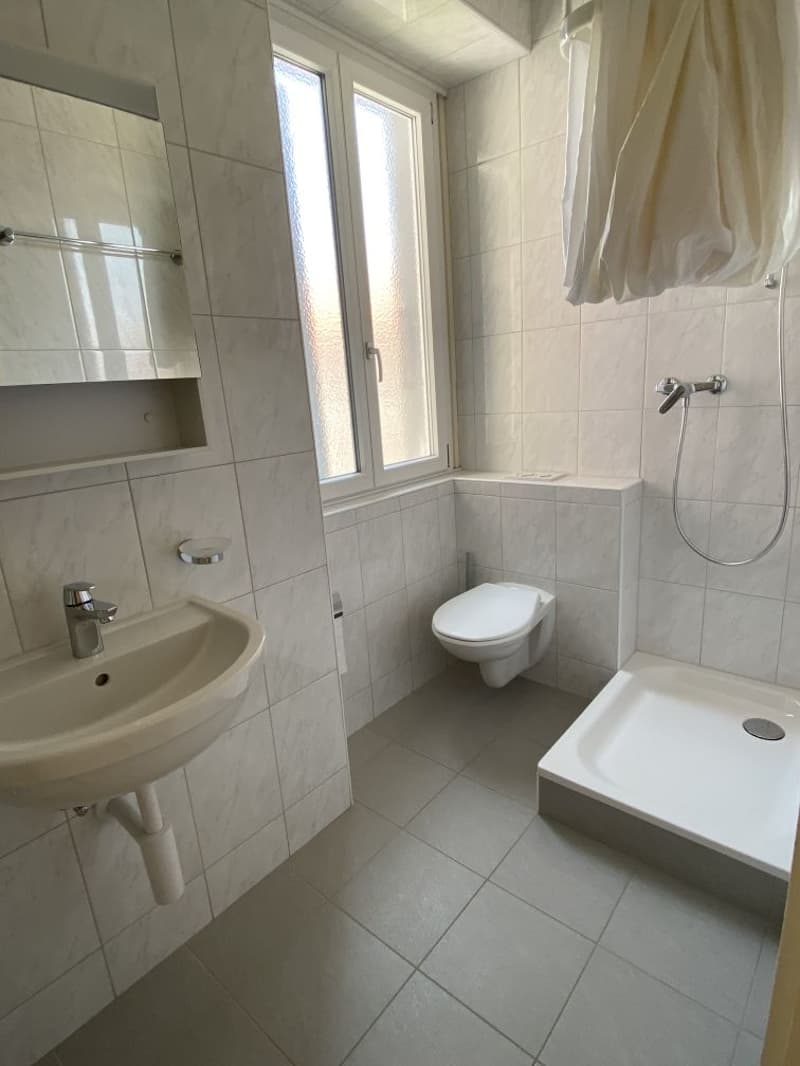 Bathroom with white tiles, sink, toilet, shower and window.