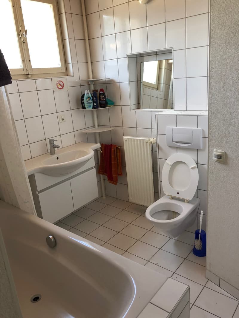A modern bathroom with white tiles, a bathtub, a sink, a toilet, and a radiator. It has a shower curtain and white cabinets. A mirror and shelves with toiletries are also present.