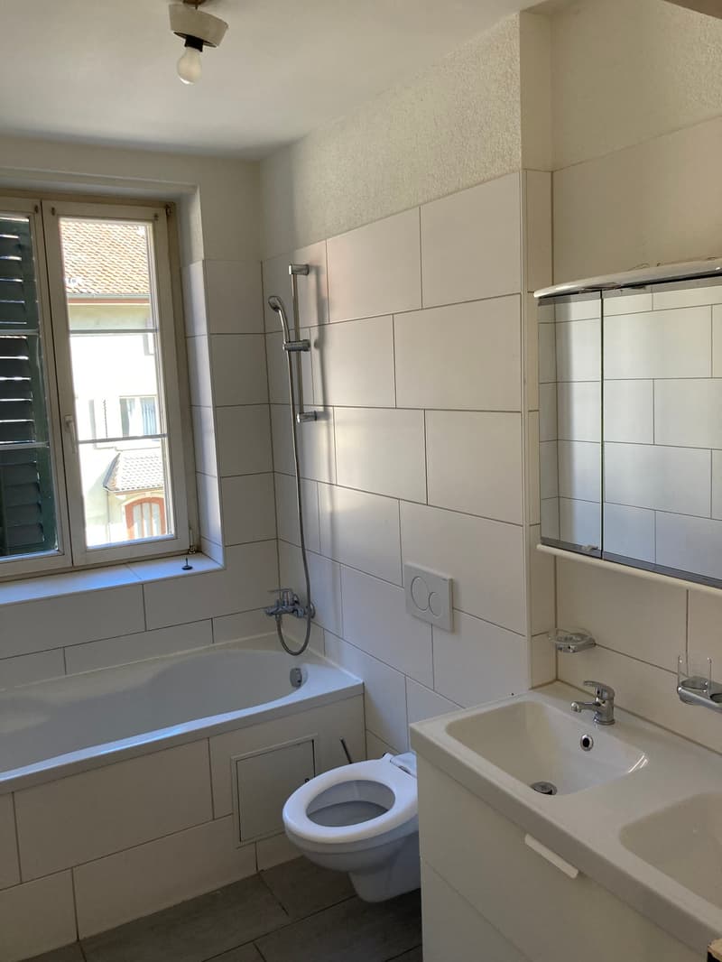 The image shows a bathroom with white tile walls, a bathtub, a toilet, and a sink. The bathroom has a window that provides natural light.