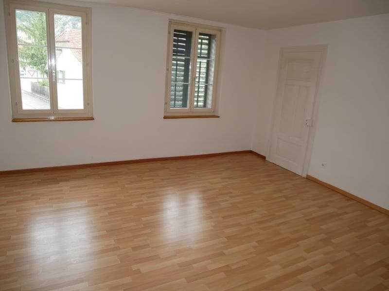 Spacious living room with hardwood flooring, large windows, and white walls. The room has a bright and airy feel with natural light coming in through the windows.
