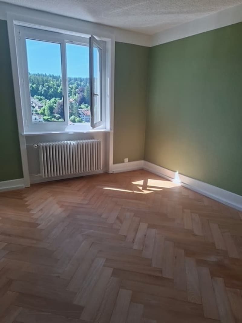 Spacious bedroom with hardwood herringbone flooring, green walls, and a large window providing a scenic view of the surrounding landscape.