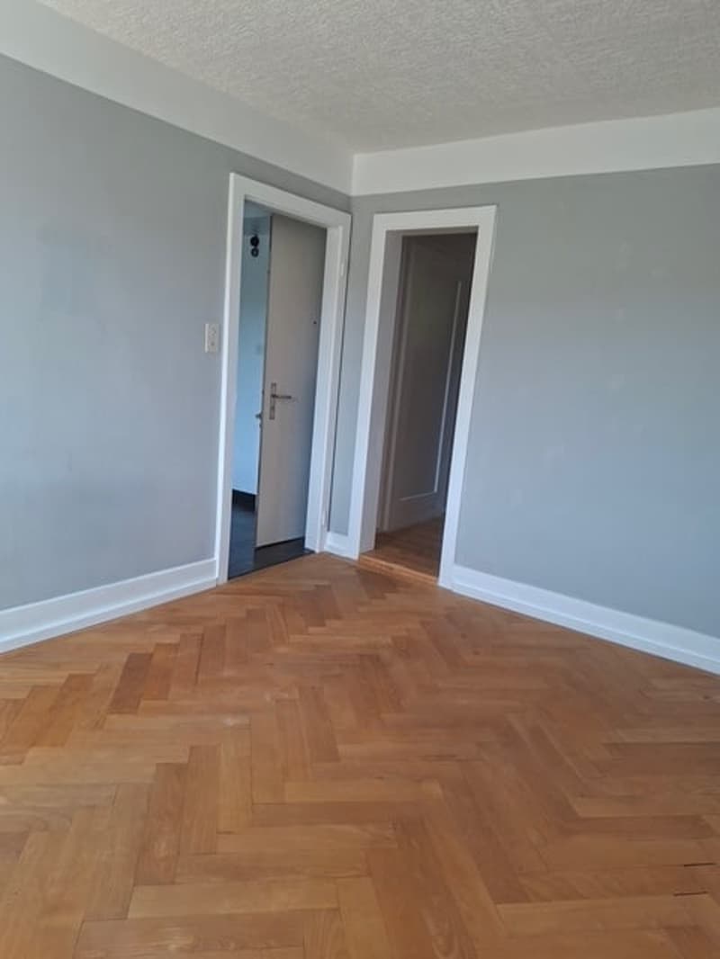 Empty room with gray walls, hardwood herringbone flooring, and two doorways visible.