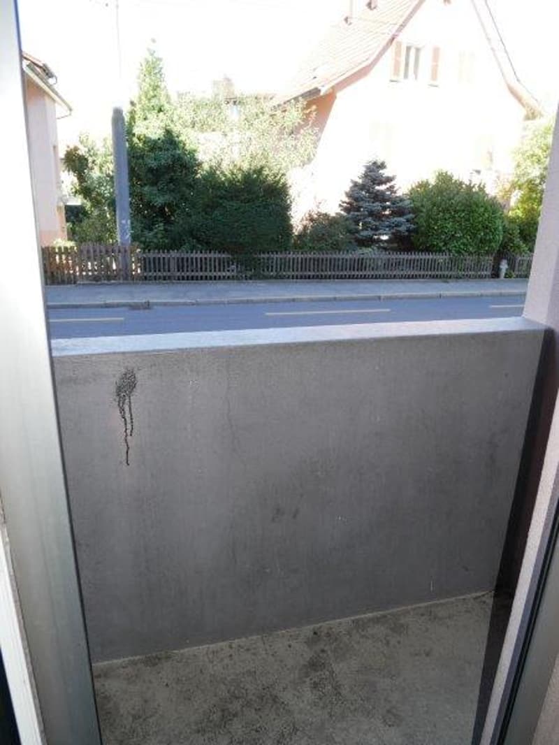 A small balcony with a concrete floor and a wall with a view of a street and a house behind a fence.