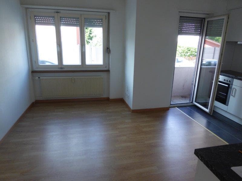 The image shows an open-plan living room and kitchen area. The room has large windows that provide natural light and access to a balcony or terrace. The floor is made of hardwood or laminate flooring. The kitchen area is equipped with modern appliances.