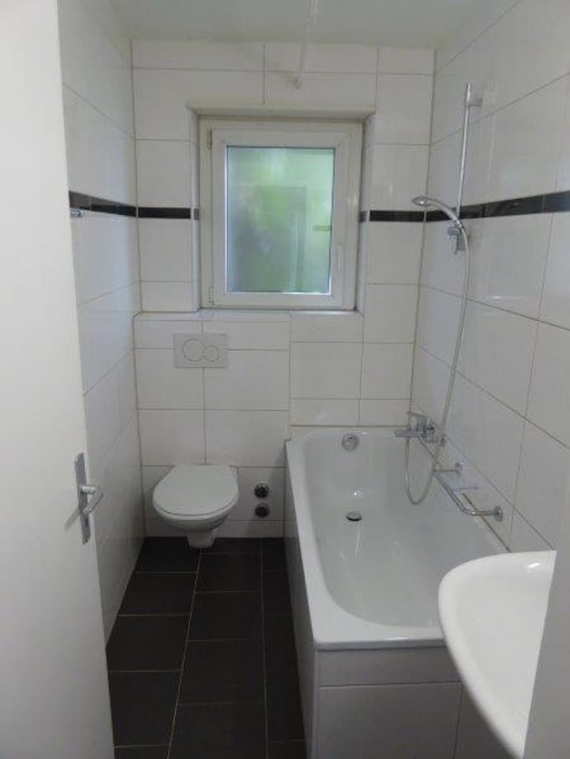 The image shows a bathroom with white tiled walls, a bathtub, a toilet, and a window. The floor is tiled in a dark color.
