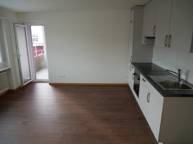 Empty kitchen, wooden floors, white walls, black countertops, stainless steel sink, built-in appliances, large windows with a balcony view.