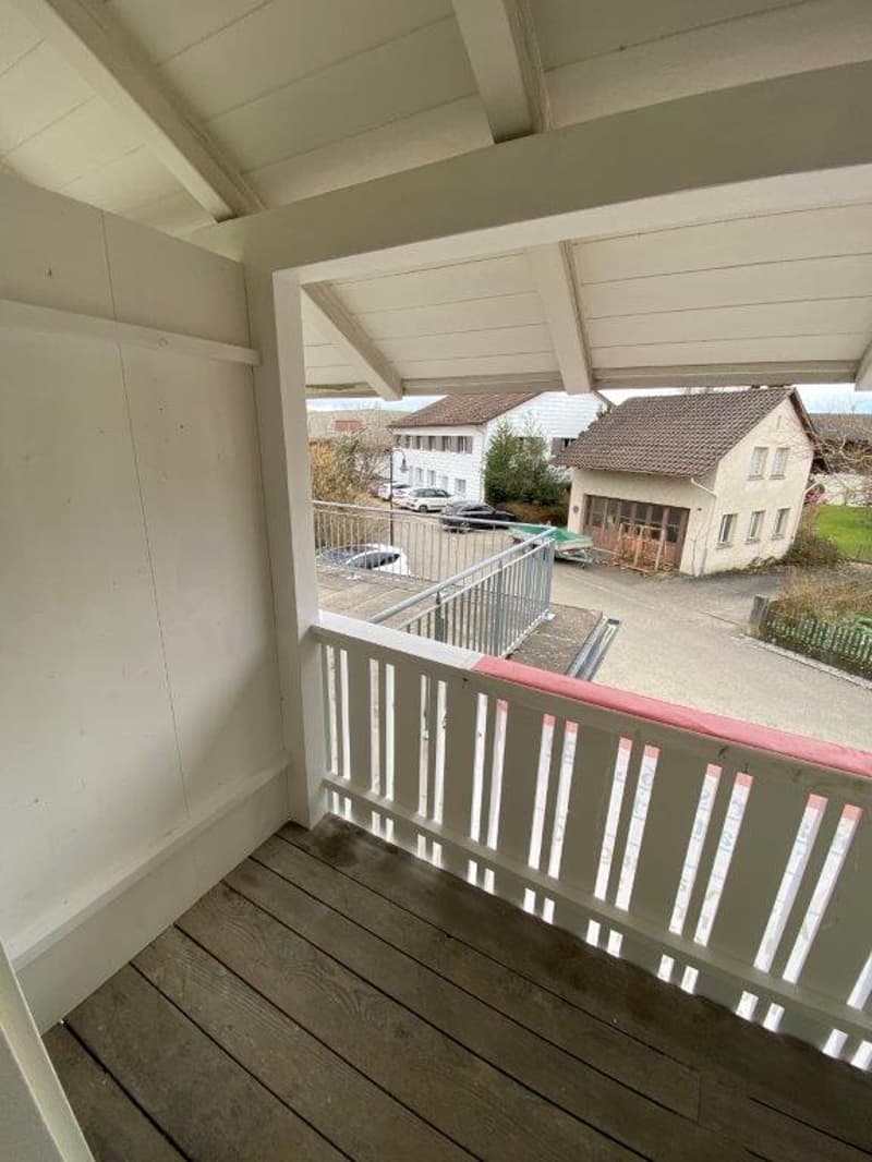 wooden floor, white walls, balcony with metal railing, view of a parking lot and houses