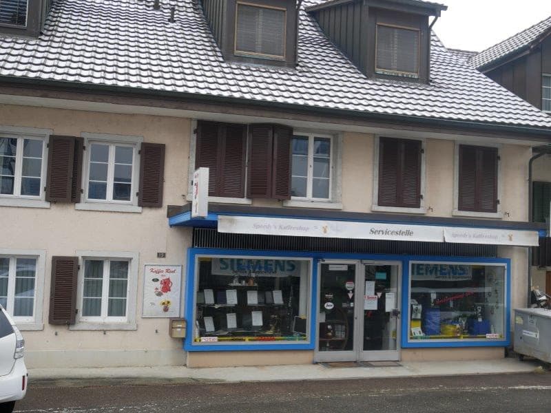 Two-story building with a tiled roof, multiple windows, and a shopfront with a sign reading 'Servicestelle'.