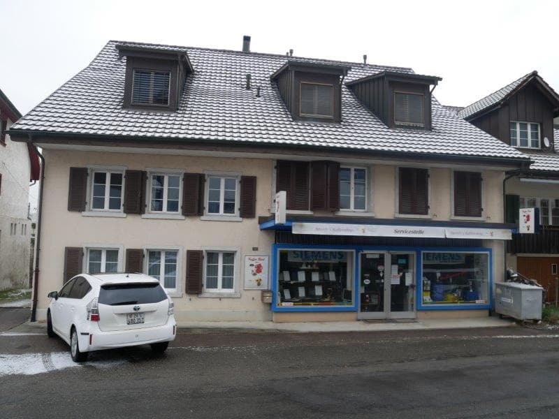 2 story house, gray roof, white windows, brown shutters, Siemens store on the ground floor, white car parked in front