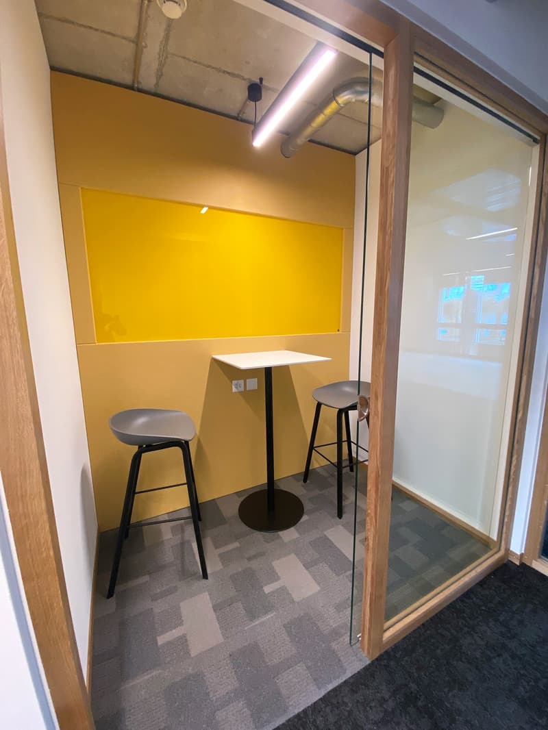 small room with a table, two stools, glass walls, yellow wall with power outlets, and ceiling lamp