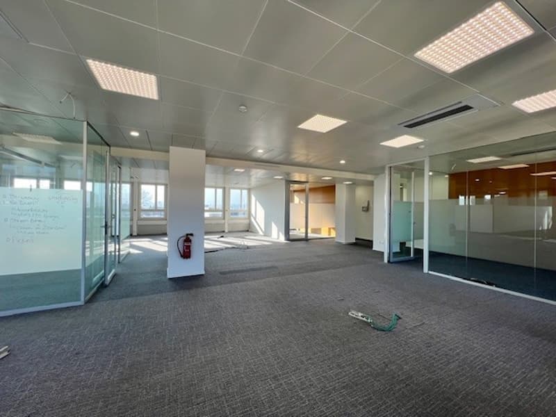 An empty office with glass walls and pillars, fire extinguisher, electrical outlets, whiteboards, gray carpet, and bright lights.